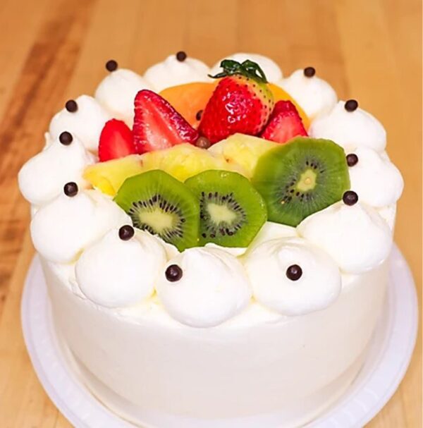 A Bombon frosted cake decorated with whipped cream dollops, chocolate beads, strawberries, kiwi slices, pineapple, and an orange slice is presented on a white plate atop a wooden surface.