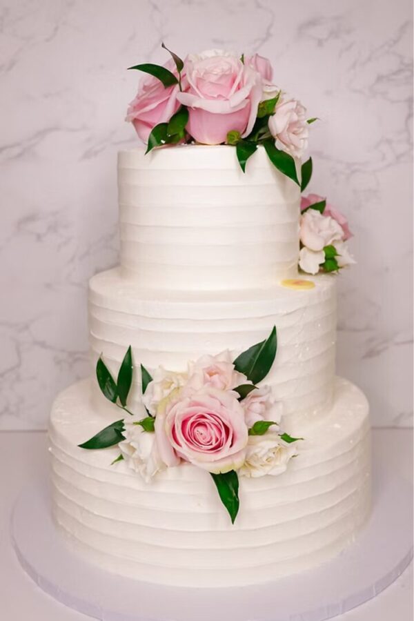 The Buttercream Roses Cake is a three-tier treat featuring textured white icing adorned with pink and white roses paired with green leaves, set against a marble backdrop.