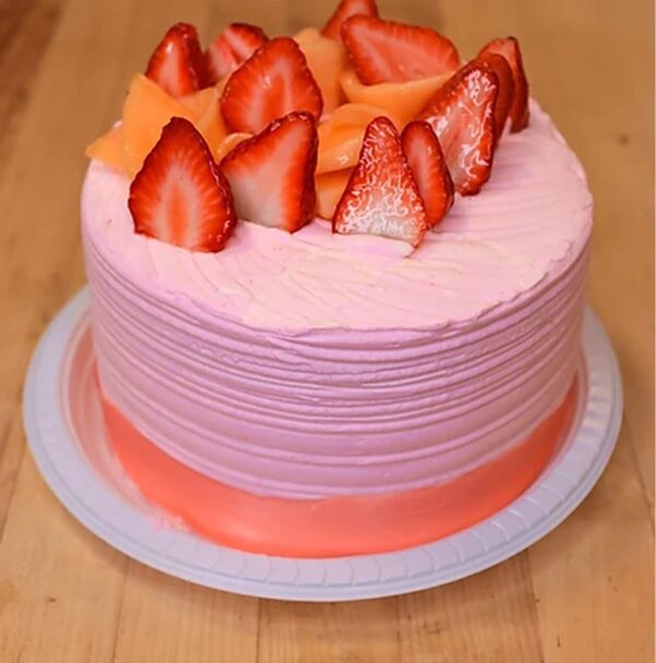 The Chupicuaro cake, adorned with pink frosting and garnished with sliced strawberries and orange fruit pieces, is elegantly presented on a white plate atop a wooden surface.