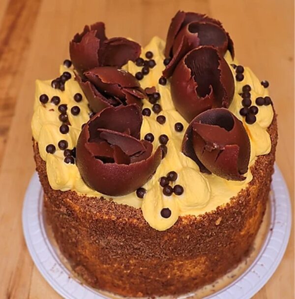 The Convento round cake features brown and yellow frosting, topped with chocolate curls and small chocolate balls, served on a white plate.