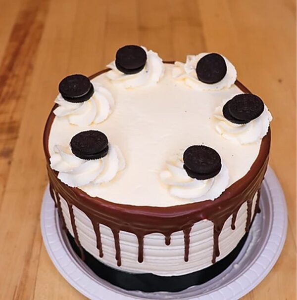 The Gabacho is a round cake on a plastic tray, featuring white frosting with chocolate drip edges. It's adorned with six cream dollops, each topped with a chocolate cookie.