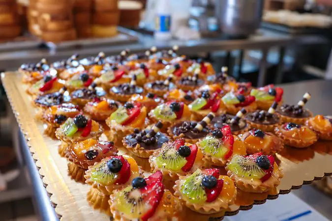 A tray of assorted fruit tarts topped with kiwi, strawberries, blueberries, and chocolate sits on a kitchen counter.