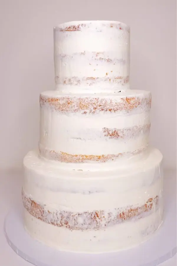The Naked Cake No Decoration features a minimalist three-tier semi-naked design with white frosting, showcasing exposed layers and a textured finish. It's placed on a simple white surface against a white backdrop.