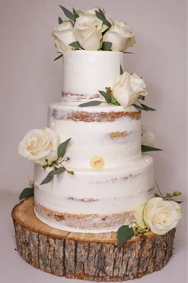 The Naked Cake With Flowers is a three-tiered semi-naked cake decorated with white roses and green leaves, resting on a wooden slice base.