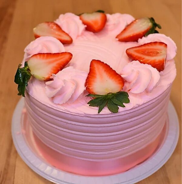 The Rosita Fresita cake, with pink frosting, sliced strawberries, and dollops of pink cream, rests on a wooden surface.