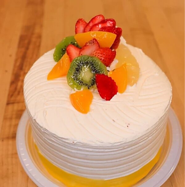 The Tutti-Futti is a frosted round cake adorned with sliced strawberries, kiwi, peaches, and orange segments, elegantly presented on a wooden surface.