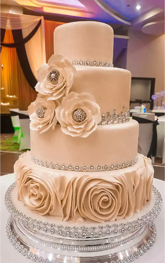 A three-tiered beige wedding cake with rose and jeweled decorations, placed on a silver stand in an elegant indoor setting.