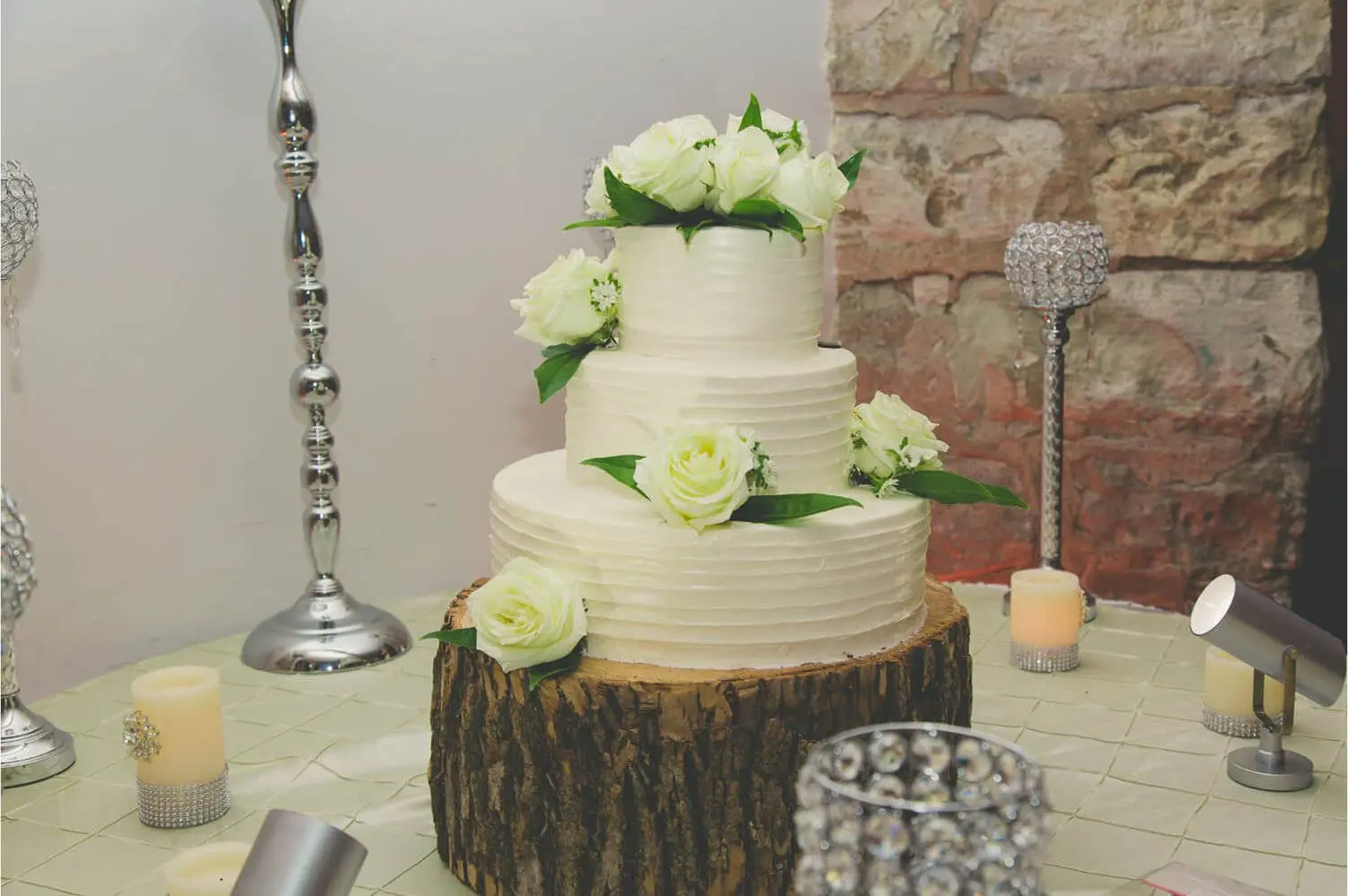 A three-tier white cake with white roses and green leaves on a wooden stand, surrounded by candles and decorative items, against a stone wall backdrop.