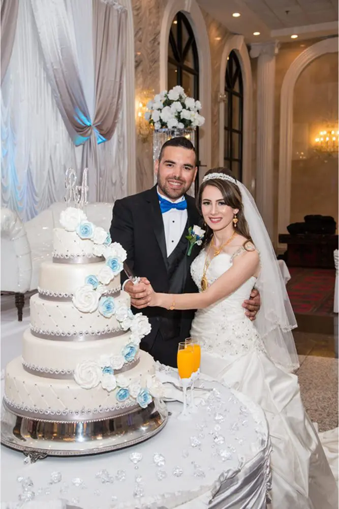A bride and groom smile while holding a cake-cutting knife. They stand beside a tiered wedding cake decorated with white and blue flowers.