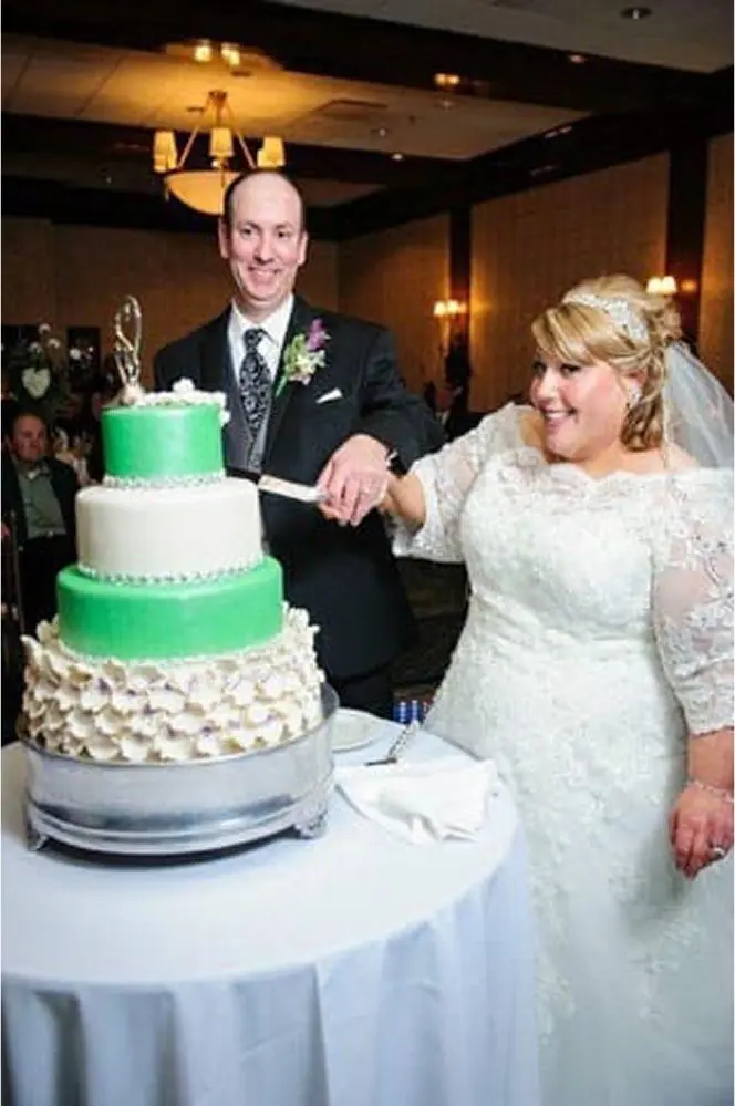 A couple cuts a three-tiered cake at their wedding reception. The cake is decorated with green and white layers and floral accents. They are standing in a banquet hall.