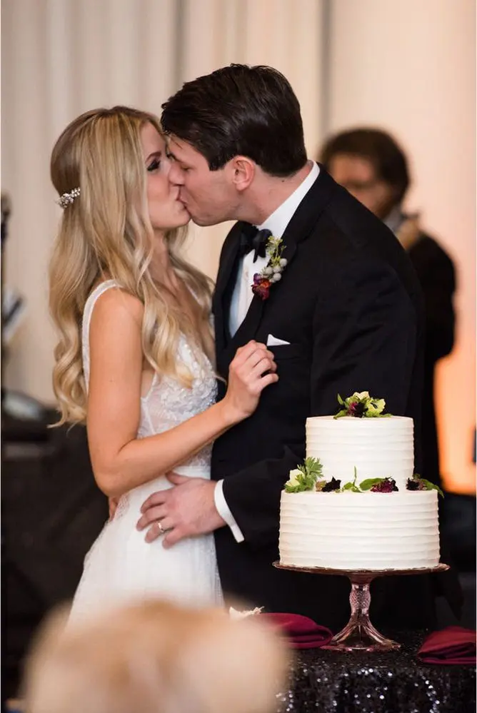 A couple in wedding attire shares a kiss beside a two-tiered white cake adorned with flowers.