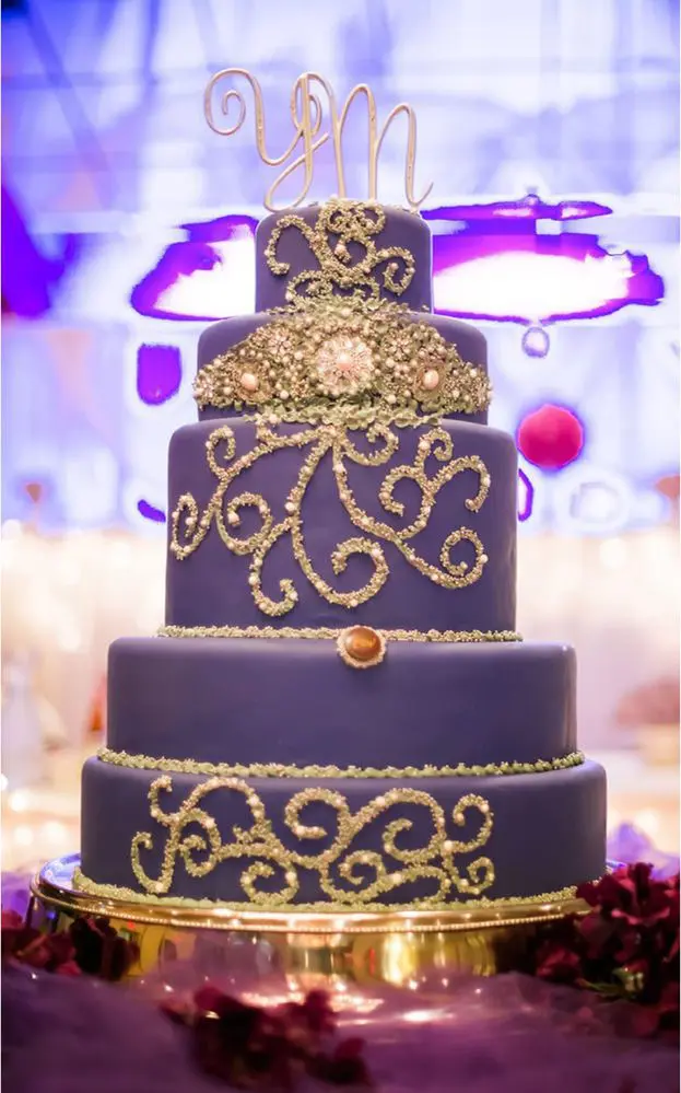 Four-tiered purple cake with gold decorative swirls and a monogram topper. Placed on a reflective base, with a blurred background of lights and pink accents.