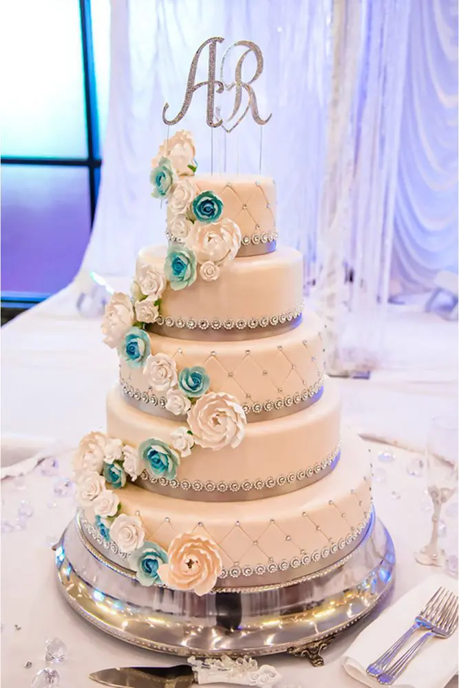 Four-tier wedding cake with white and blue roses, silver decorative accents, and a monogram topper "AR.