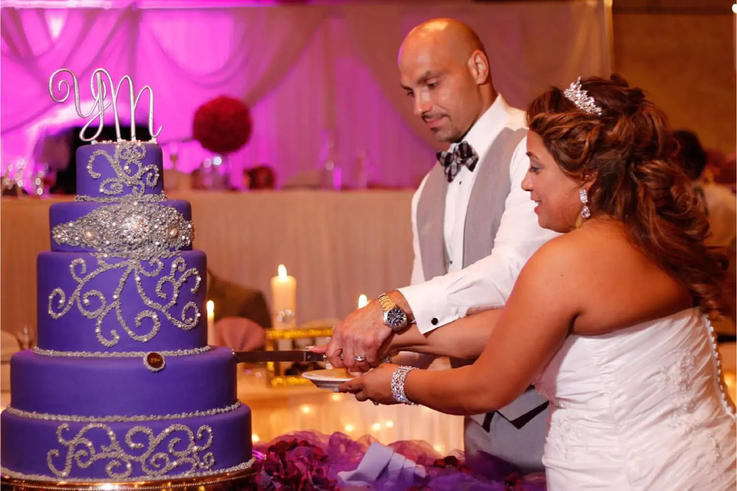 A couple in wedding attire cuts a large, ornate purple cake decorated with silver details and topped with initials in a warmly lit reception setting.