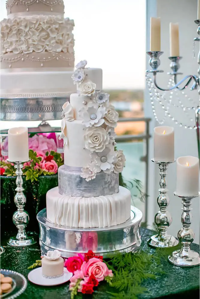 Three-tier white cake with floral decorations and silver accents on a silver stand, surrounded by lit candles and pink flowers.