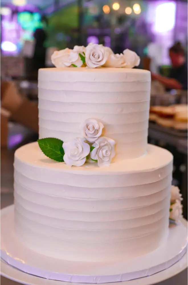 Two-tiered white cake with smooth icing and white rose decorations, placed on a white cake board.
