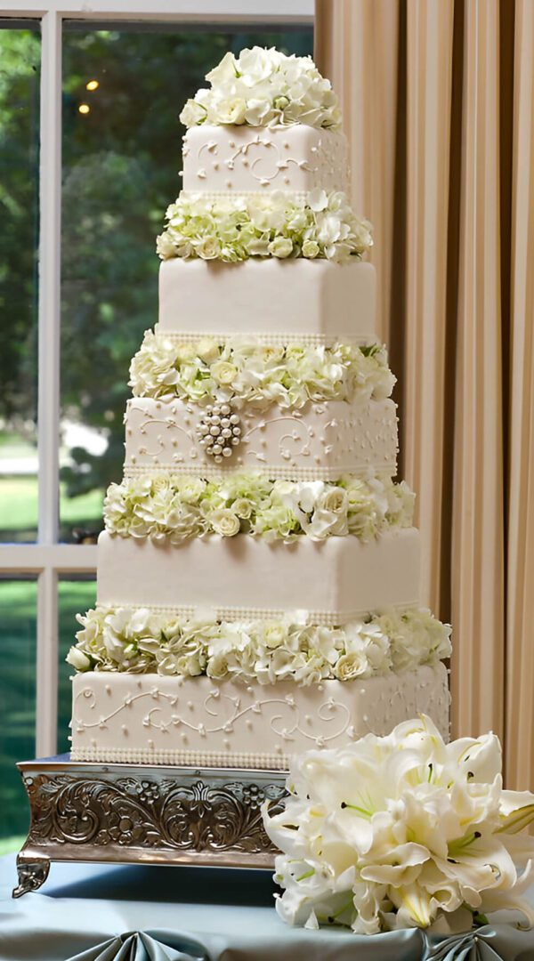 A tall, tiered white wedding cake adorned with white flowers and decorative frosting details, placed on an ornate silver stand.