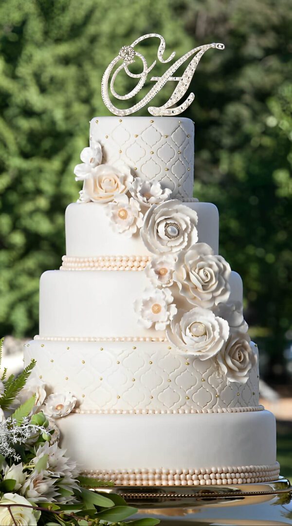A tiered white wedding cake with quilted patterns, adorned with white roses and pearl-like beads. The top features a decorative letter "A." Greenery surrounds the base.