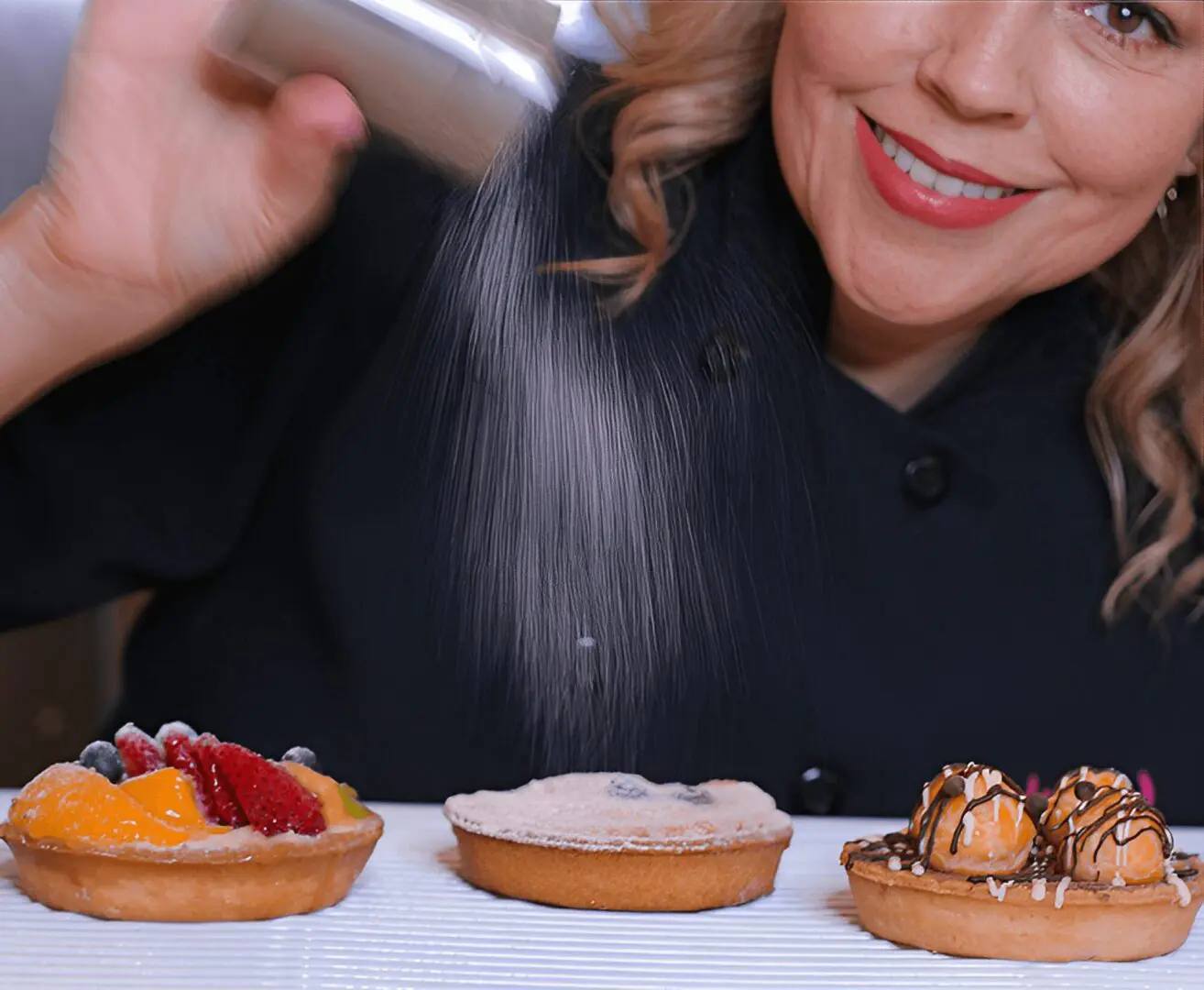A person smiling while sprinkling powdered sugar over three pies, topped with assorted fruits and toppings, on a table.